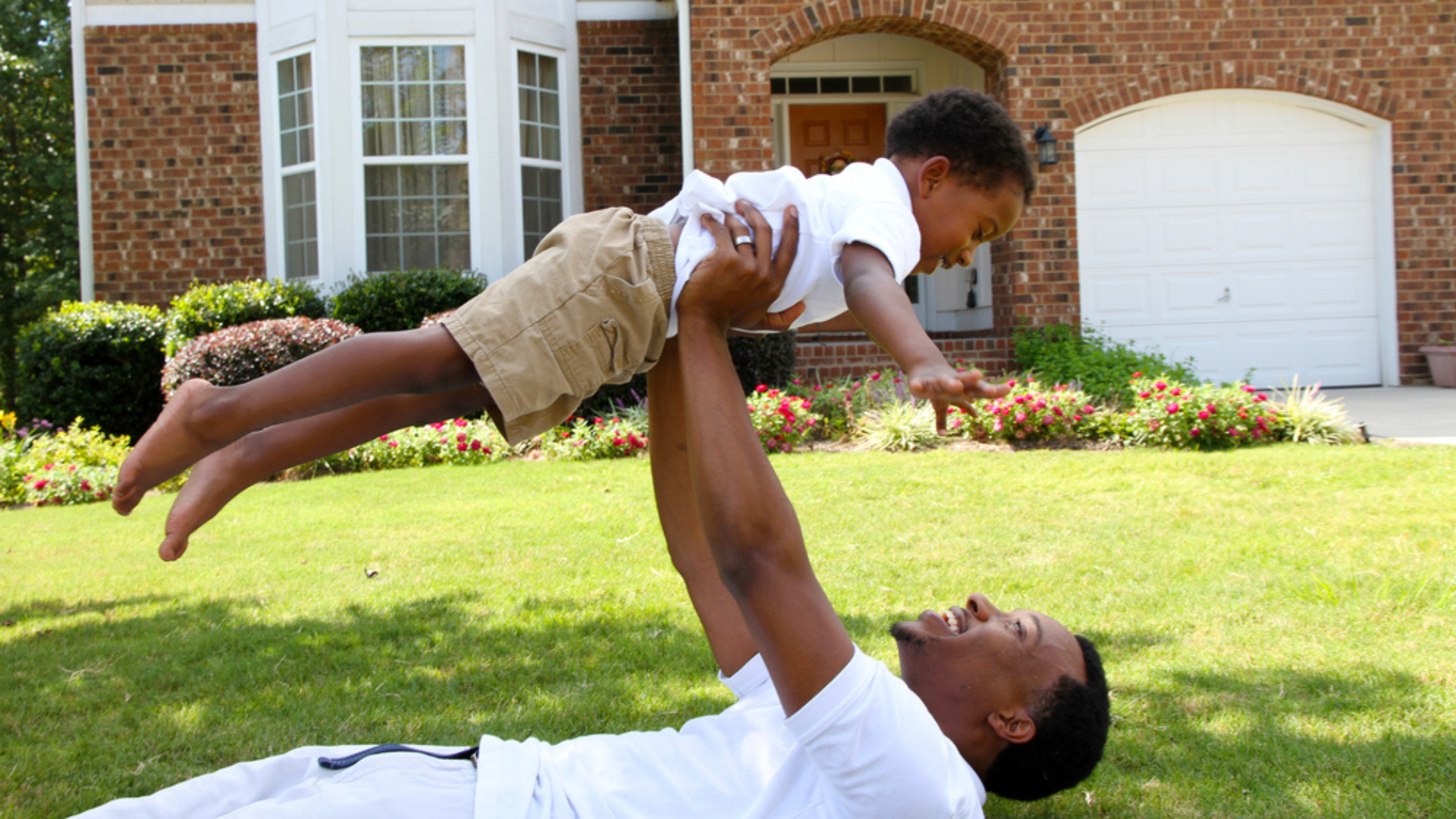 Dad lifting child while laying on grass in front of a brick home